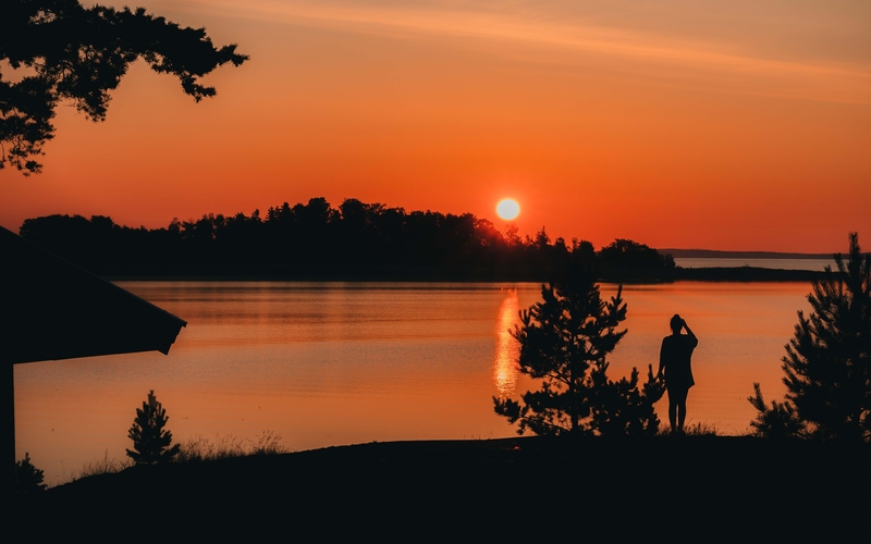 Solnedgång över åländska berg och vatten om sommaren, silhuett av en kvinna i förgrunden till höger.